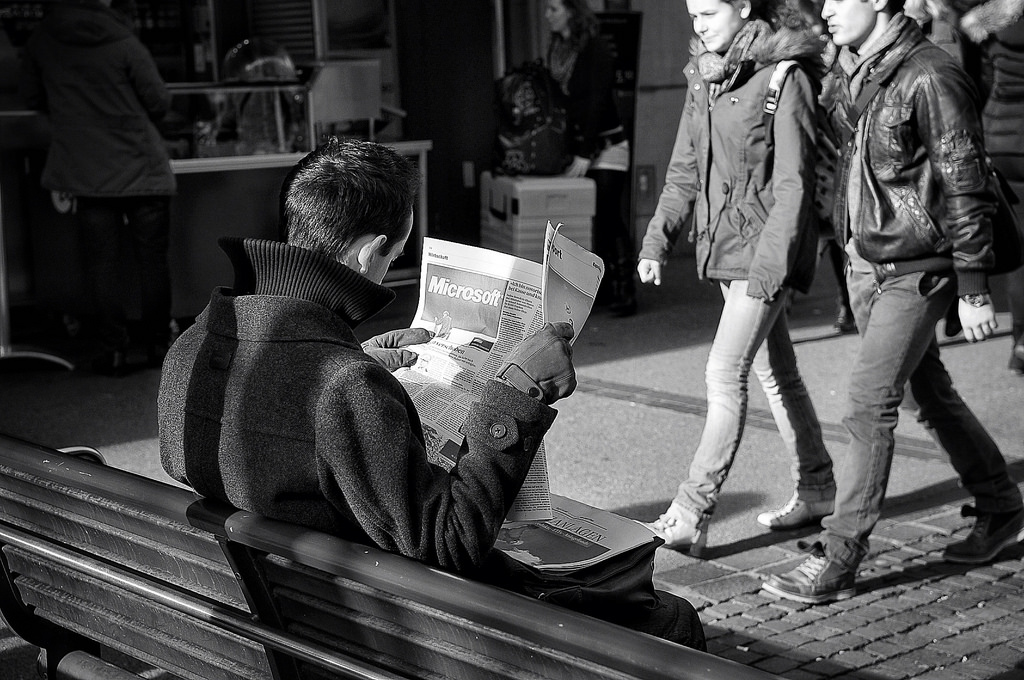 Man sitting on park bench on street reading newspaper with pronounced Microsoft ad visible.