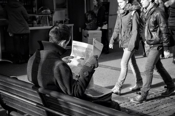 Man sitting on park bench on street reading newspaper with pronounced Microsoft ad visible.