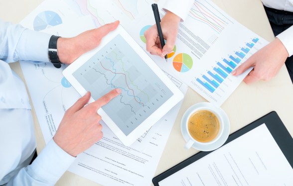 Two business people (just see their hands) workign on charts and graphs on paper and a tablet.