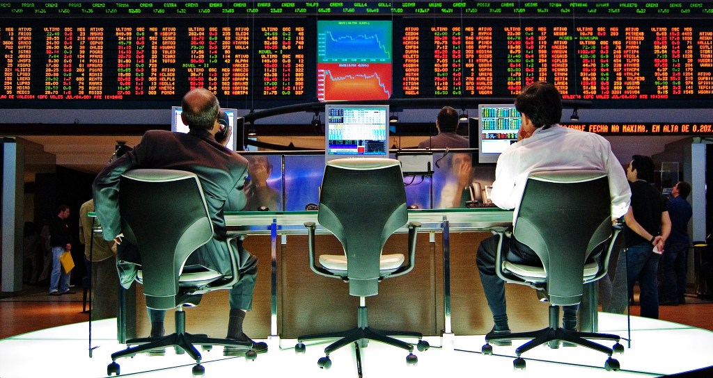 Two men sitting in front of bank of monitors with stock prices.