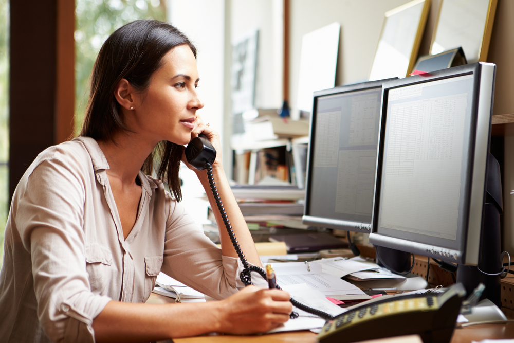 Woman on phone in front of two big computer screens taking notes.
