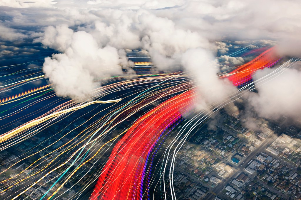 Streaks of colored lights race beneath the clouds and over an urban scene in a metaphor for cloud computing and communications technology.