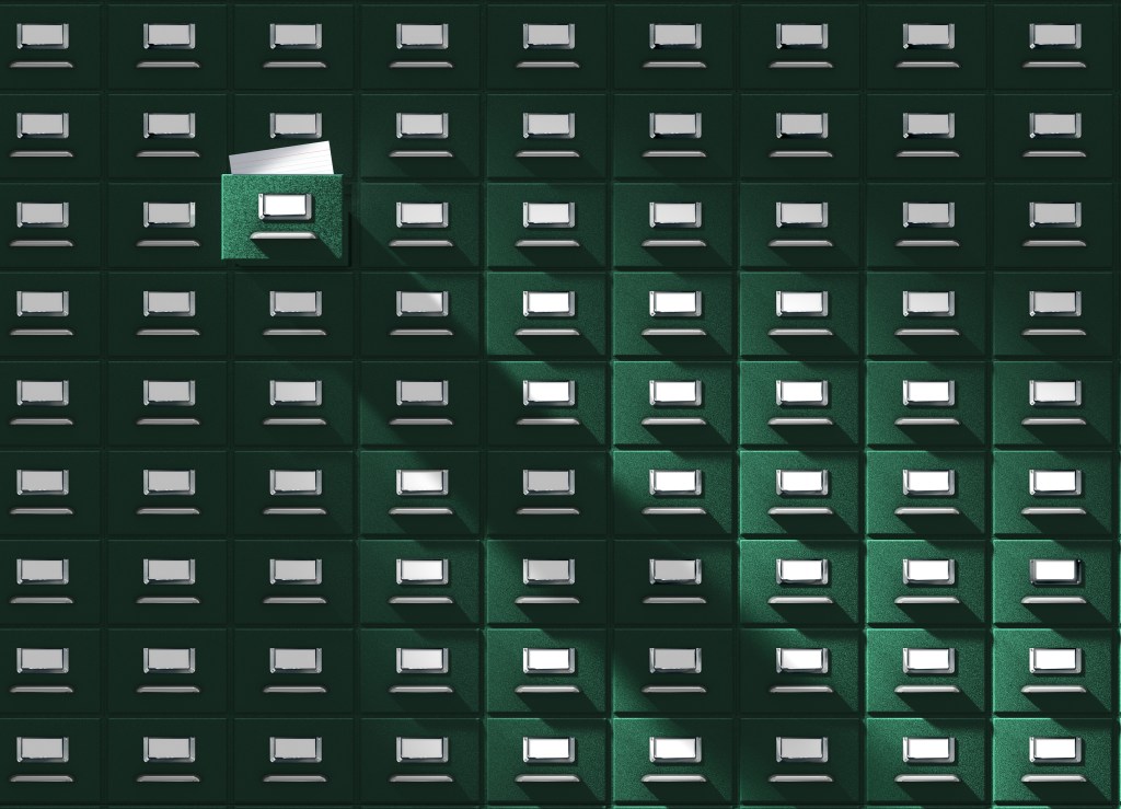 An immense card catalog system with one drawer pulled out, revealing a blank catalog card. This vintage database is floodlighted from the upper left side, giving it a mysterious edge.