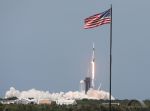 A SpaceX Falcon 9 rocket lifts off, framed by an American flag atop a flagpole.