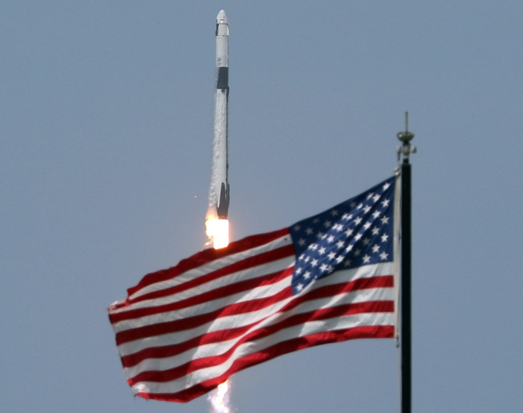 SpaceX rocket lifts off with American flag fluttering in the foreground