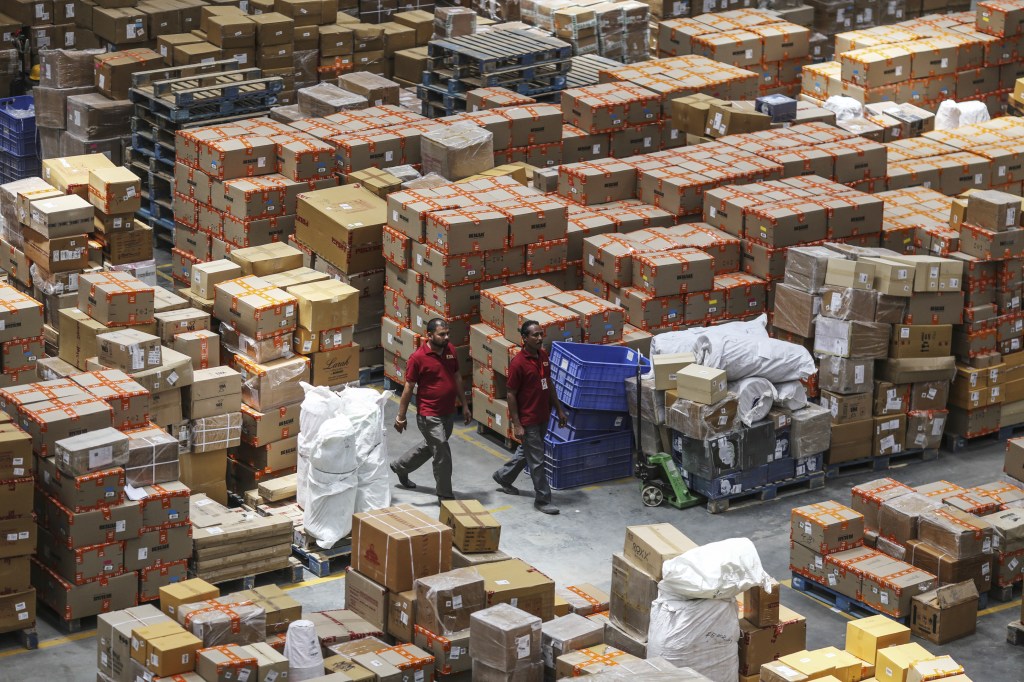 Employees walk past boxes stacked on pallets at a warehouse