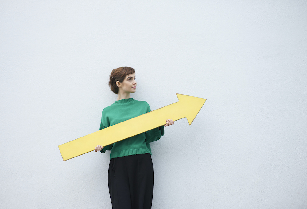A young student woman stands against a white wall holding a large yellow arrow sign.