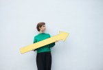 A young student woman stands against a white wall holding a large yellow arrow sign.
