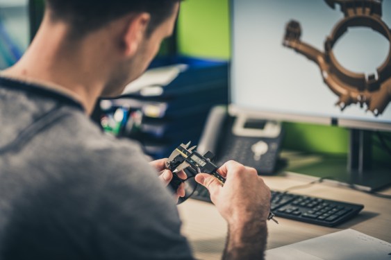 Over-the-shoulder view of an engineer measuring a machine part while working on a 3D model of it.