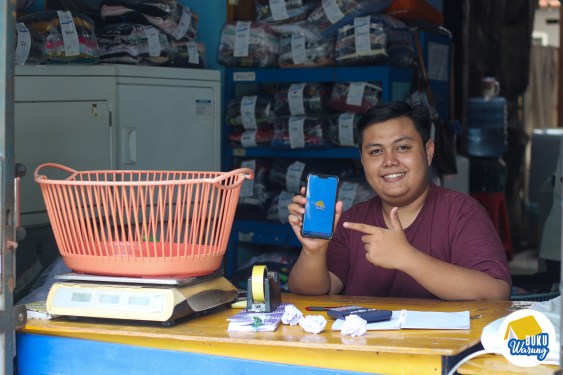 An image of an Indonesian merchant holding a smartphone with BukuWarung's app