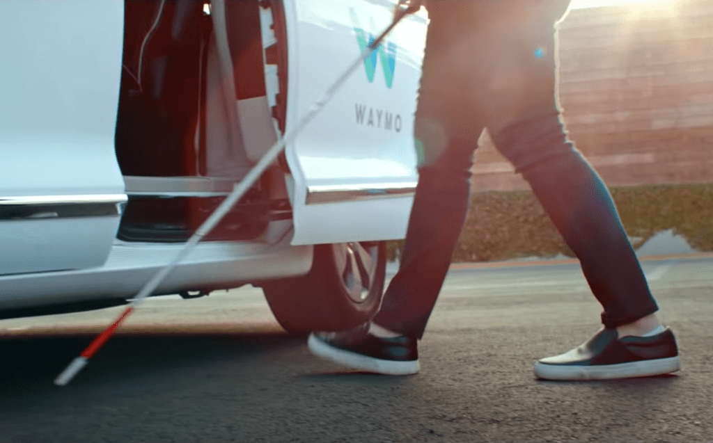 Image shows legs and cane of a visually impaired person as they prepare to enter the open door of a Waymo self-driving vehicle.