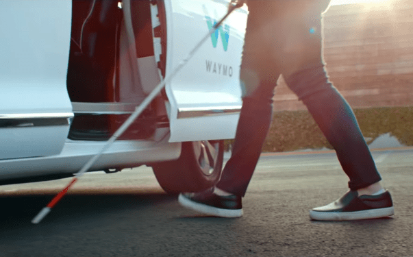 Image shows legs and cane of a visually impaired person as they prepare to enter the open door of a Waymo self-driving vehicle.