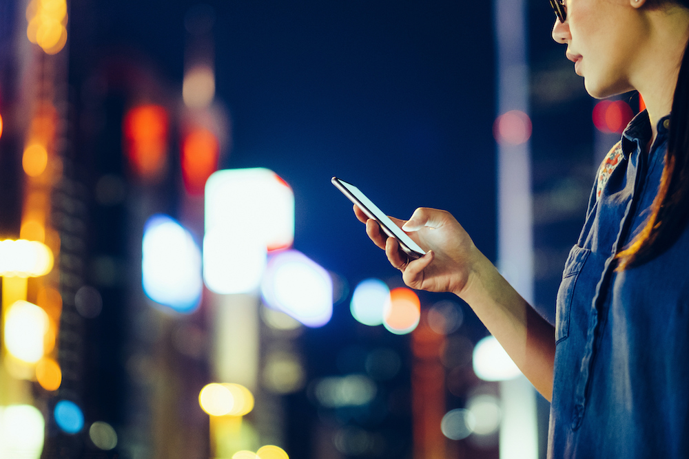 Cropped image of young woman using mobile phone outdoors at night, against city buildings with multi coloured neon lights