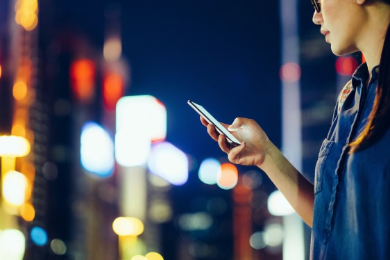 Cropped image of young woman using mobile phone outdoors at night, against city buildings with multi coloured neon lights