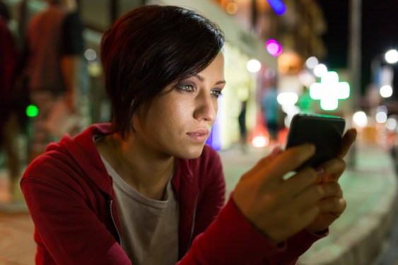 Side view of young woman using smart phone on street by night. Wears casual clothes. Street view and lights on background.