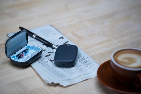 Whisper hearing aid box on newspaper with pen and crossword puzzle. Whisper Brain is sitting on newspaper. Cup of Capuccino is next to it to show relative size.
