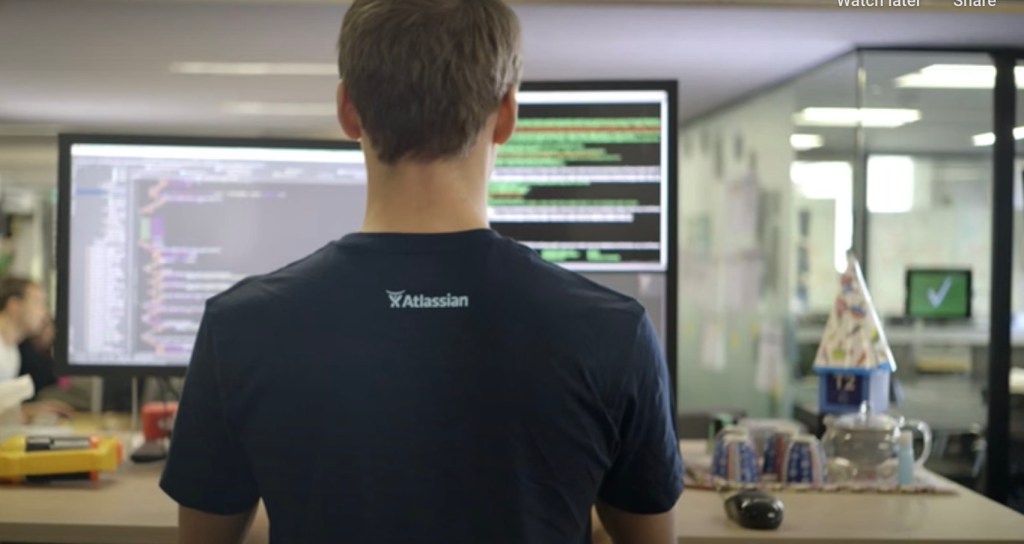 Programmer working in front of large monitors viewed from behind. He is wearing an Atlassian T-shirt.