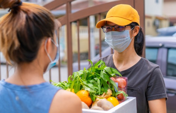 A grocery store delivery person wearing an orange cap and face mask. She is delivering groceries to a woman.