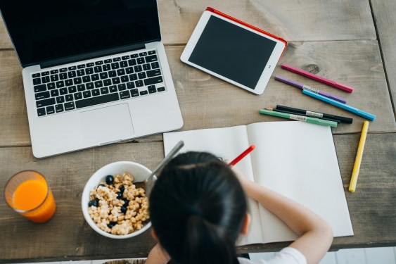 Overhead view of girl having breakfast while attending online school classes with laptop and digital tablet.