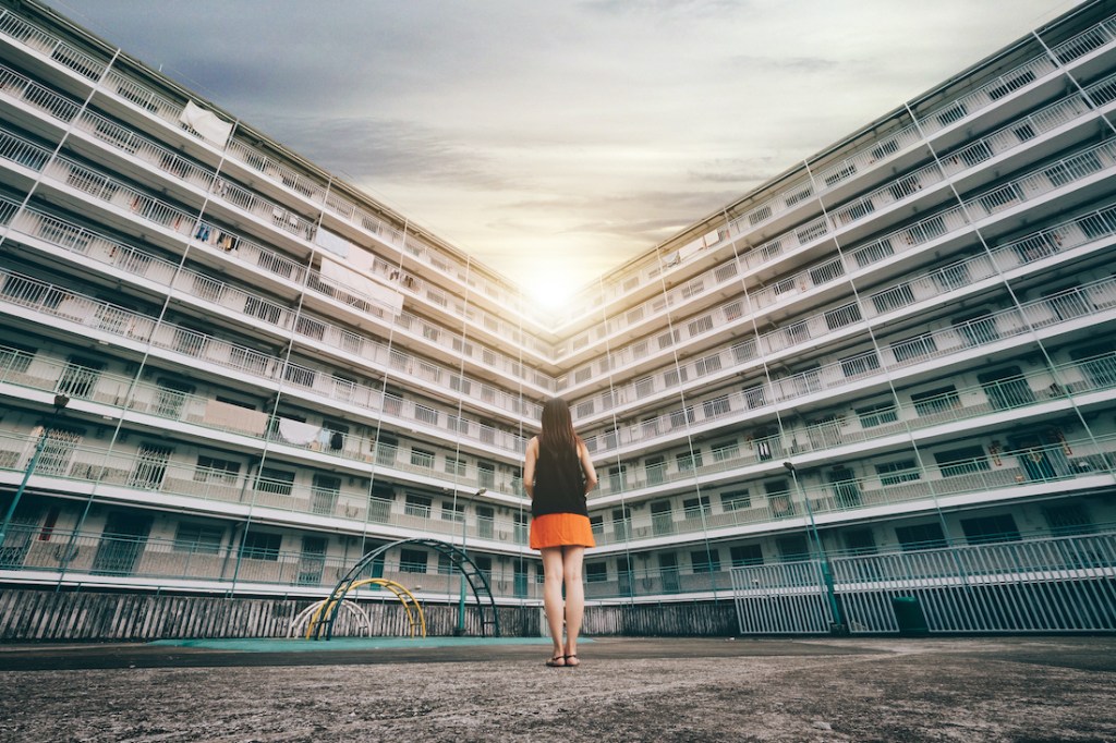 The rear view of woman standing against old apartment building, looking towards the shining sun.