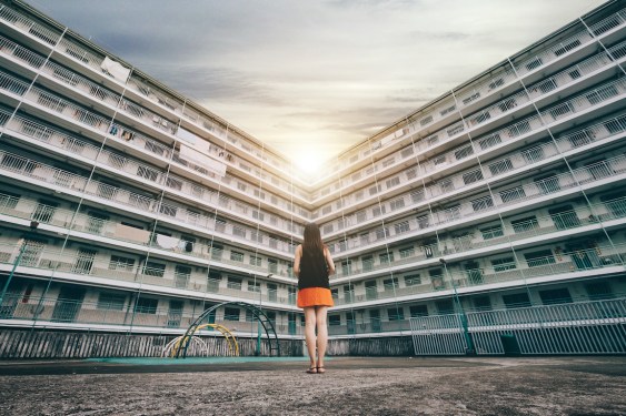 The rear view of woman standing against old apartment building, looking towards the shining sun.