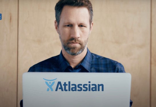Bearded man sitting in front of laptop with Atlassian sticker on it.