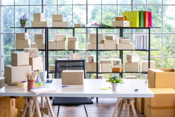 A photo of a laptop on a desk, surrounded by shelves full of packages in front of a large window