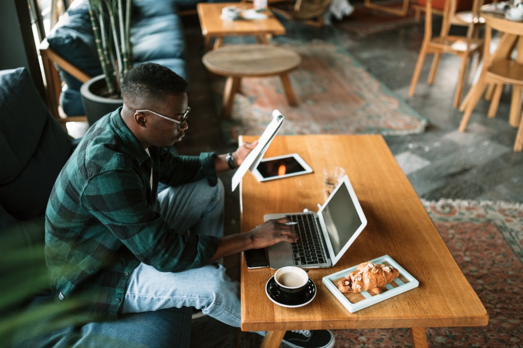 Side view of a young African American male worker sitting in a cafe, casually working using his laptop
