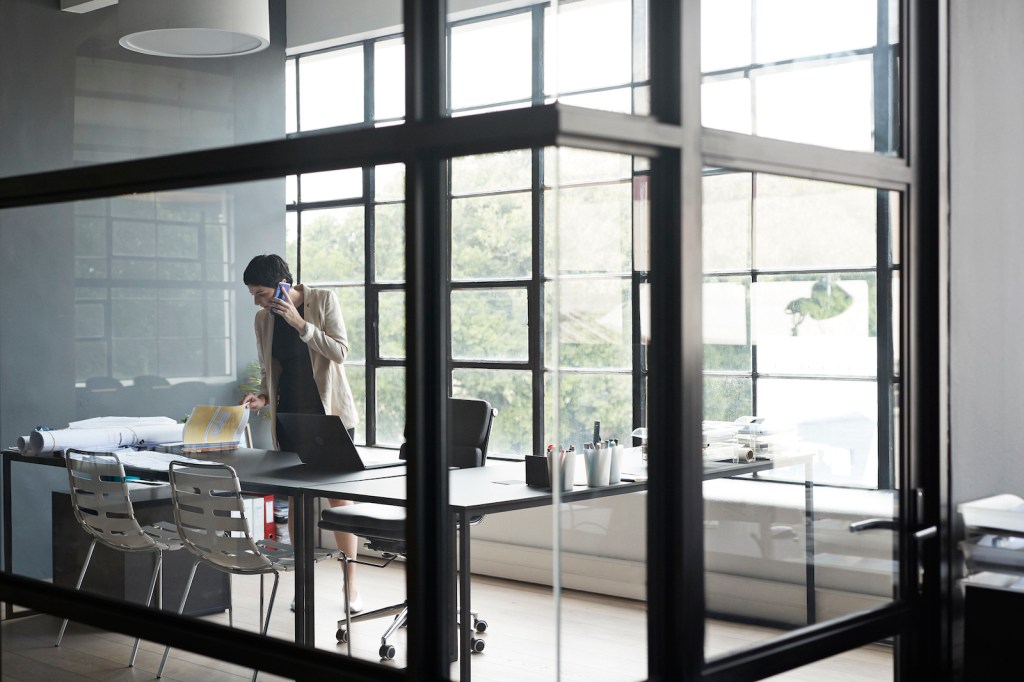 Businesswoman talking through mobile phone in modern office seen through glass wall