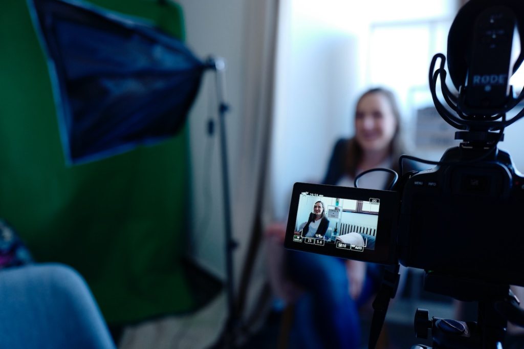 A woman is sitting in front of a video camera and some lighting gear.