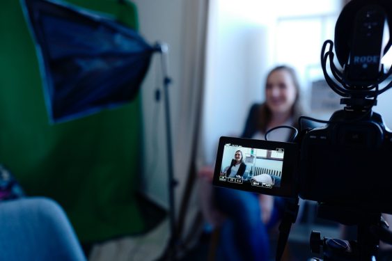 A woman is sitting in front of a video camera and some lighting gear.