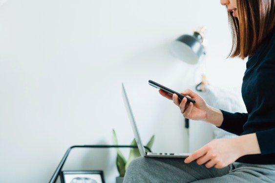 Side profile of young Asian woman working from home, working on a laptop and using smartphone in bed