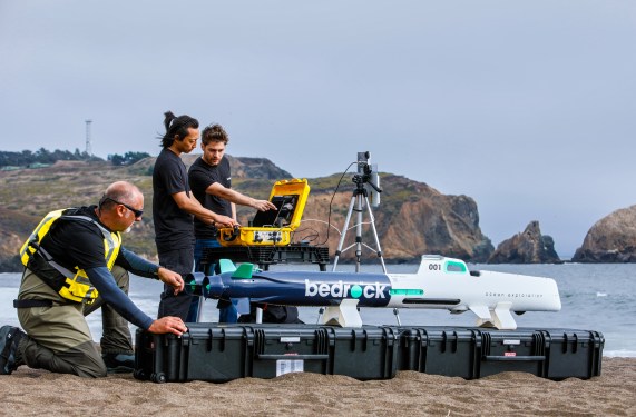 Bedrock cofounders on a beach next to their small autonomous underwater vehicle.