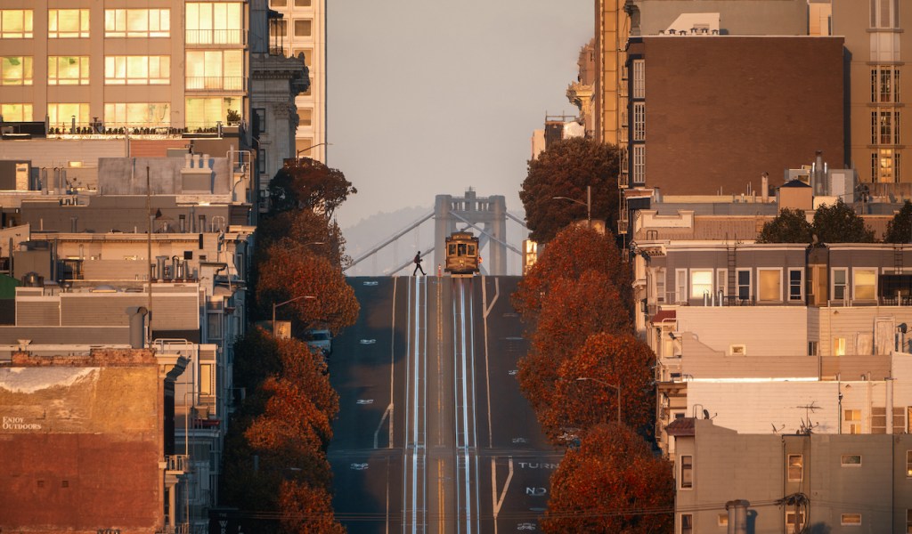 A cable car at the top of a hill in San Francisco with the Bay Bridge in the background.