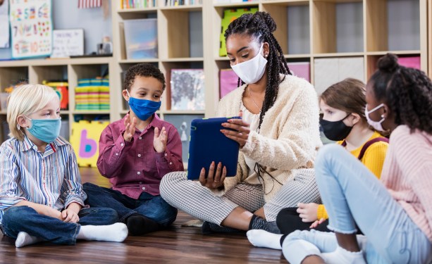 Image of a teacher sitting on the floor with young students and a tablet in a classroom.