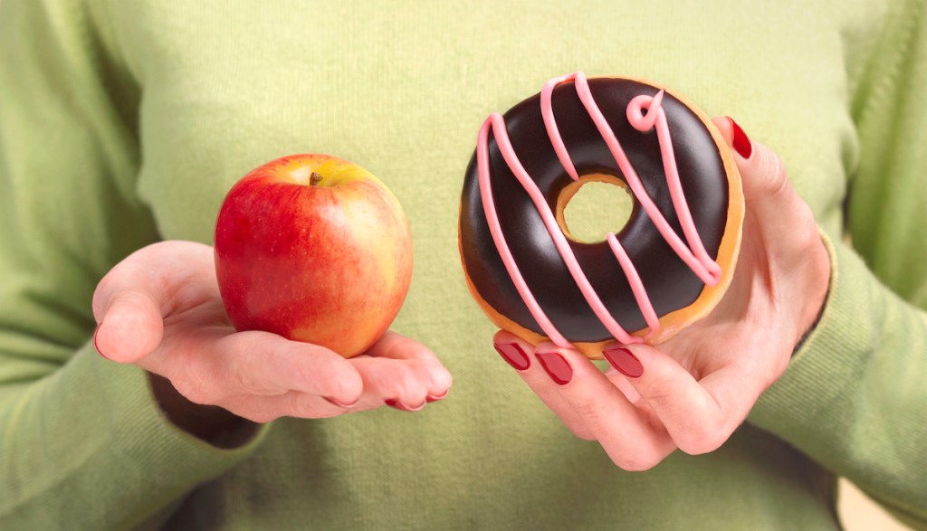 A close up of a woman's hands, one holding an apple the other hand holding a doughnut