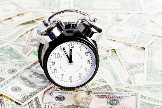 A pile of US currency, topped with an old-fashioned alarm clock reminds us that time is money and very valuable. As the clock shows just before12, could also indicate last-minute purchases or payments.