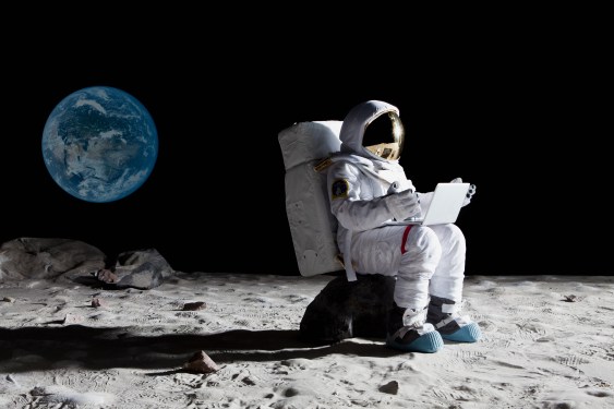 Image of an astronaut on the moon sitting on a rock using a laptop.