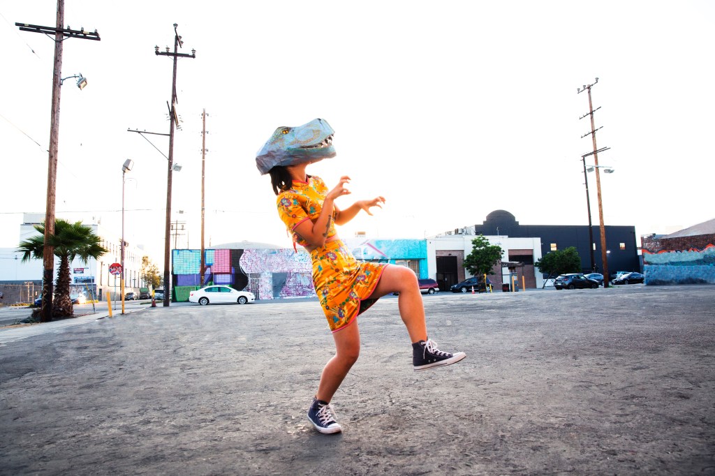 Image of a young woman wearing a handmade dinosaur mask in an empty parking lot.