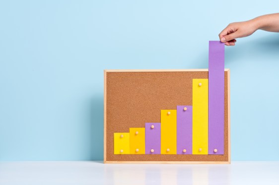 Hand Pulling Up Rising Bar Chart on Cork Board Against Light Blue Colored Background.