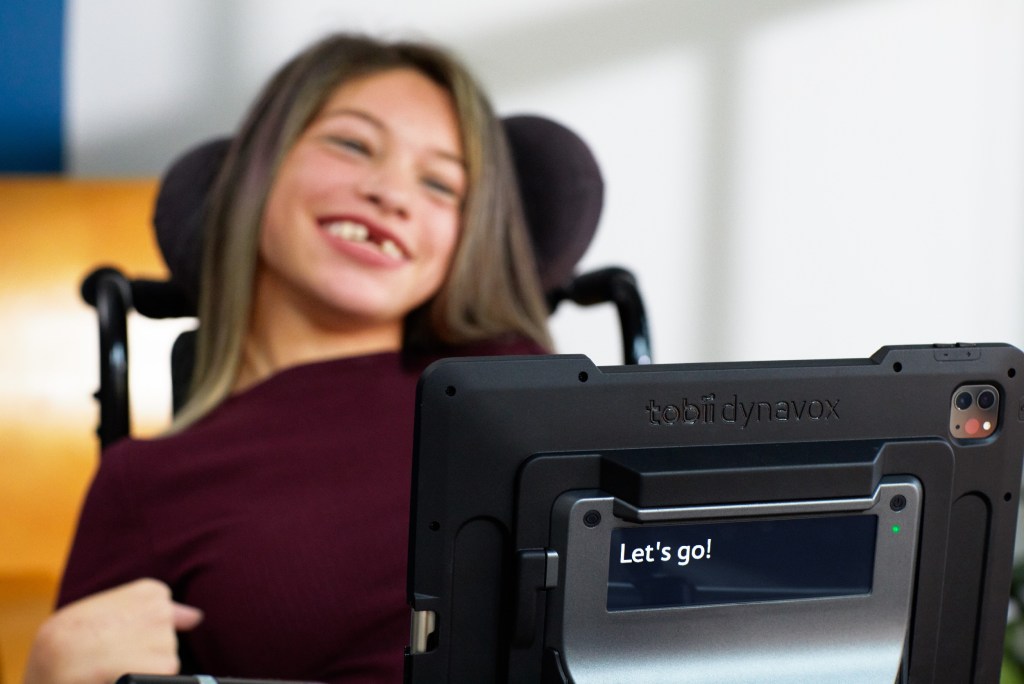 A smiling woman in a wheelchair uses an eye-tracking system on an iPad.