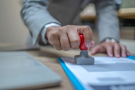 Close-up Of A Person's Hand Stamping With Approved Stamp On Document At Desk