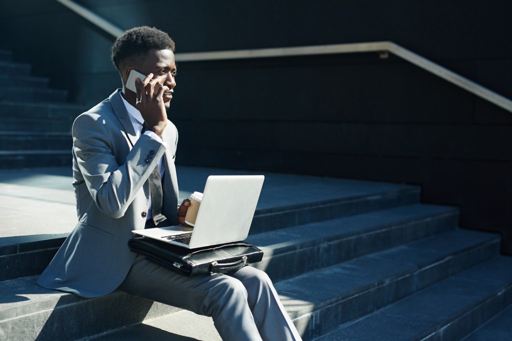 Sales person sitting outside on steps with coffee and laptop making sales calls on smartphone.