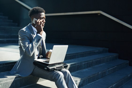 Sales person sitting outside on steps with coffee and laptop making sales calls on smartphone.