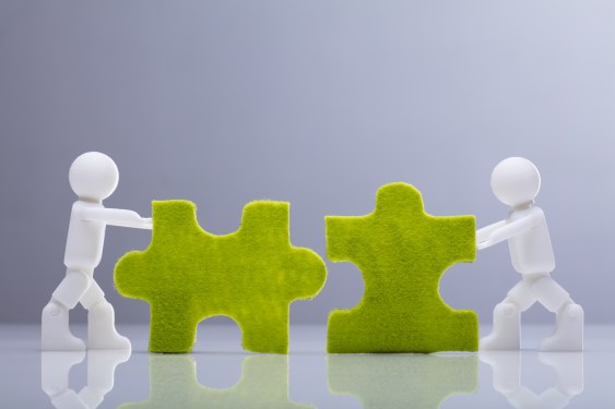 Side View Of Two Miniature Human Figures Solving Green Jigsaw Puzzles On Grey Background