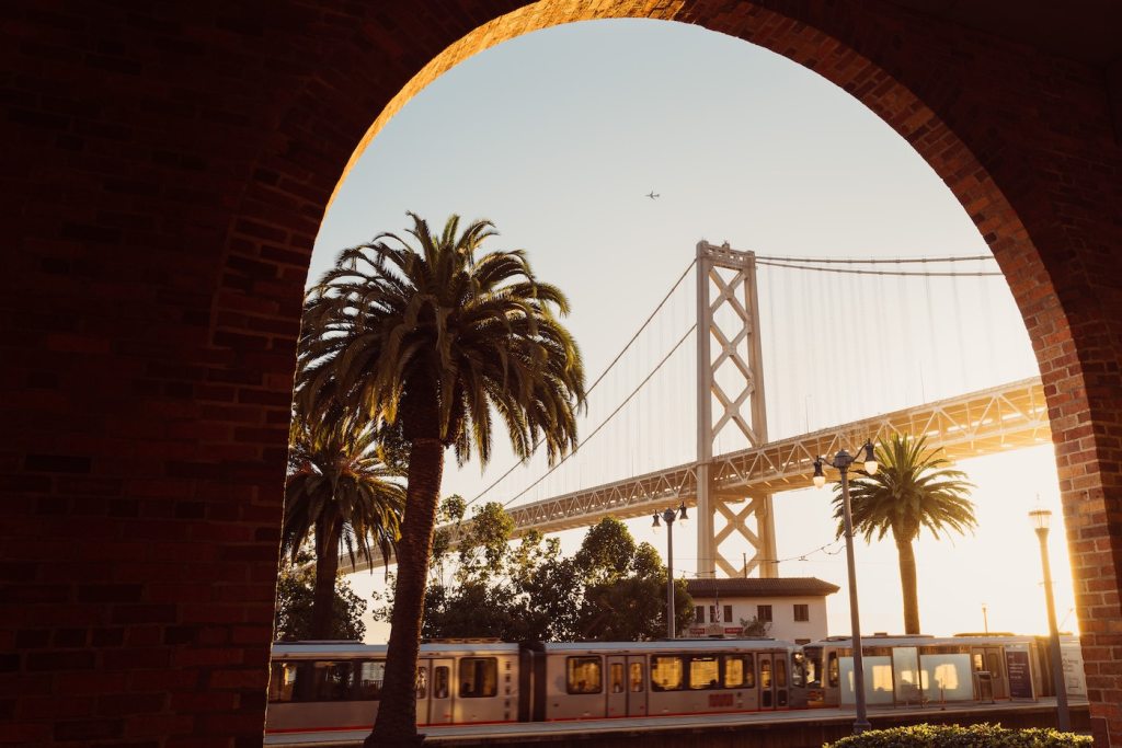 A photo of the San Francisco-Oakland Bay Bridge as seen through an archway with a ligh-rail vehicle in the background.