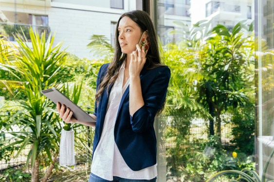 Woman with digital tablet and face mask in hand talking on the phone while standing by window in garden, Cologne, NRW, Germany