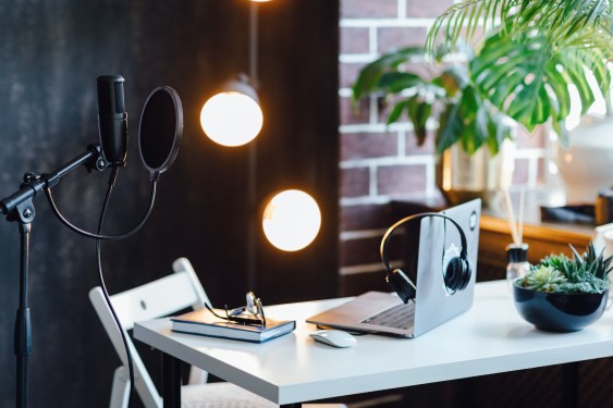 Podcast streaming at home. Audio studio with laptop, microphone with pop filter and headphones on white table against black wall with warm lights. Blogger concept.