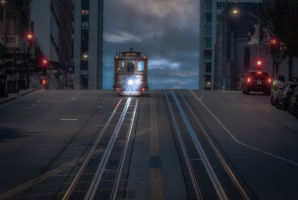 A cable car with its headlight illuminated descends a hill in San Francisco, CA
