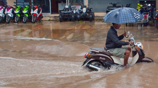 Man rides scooter thorugh flooding, while holding up an umbrella